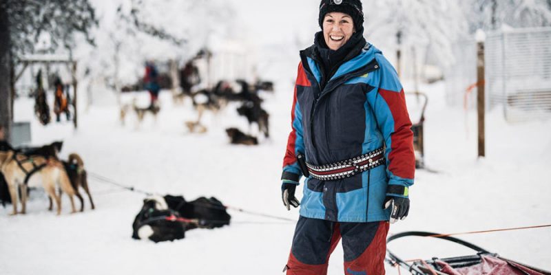 Frau im Schneeanzug steht vor Huskyfarm im Schnee